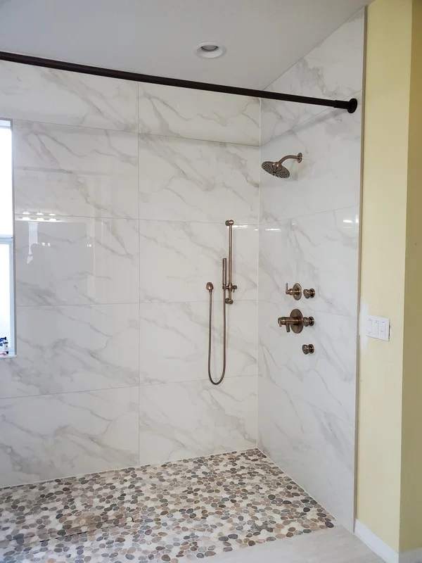 Walk-in shower installation with marble-style wall tile, pebble shower floor, rainfall showerhead, handheld sprayer, and modern glass shower enclosure for a clean bathroom upgrade.